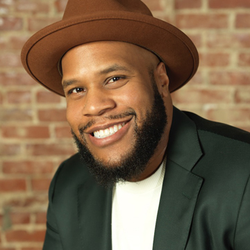 Smiling man with a beard wears a brown hat, dark blazer, and white shirt, posing in front of a brick wall background, exuding confidence and success as if ready to talk about money matters.