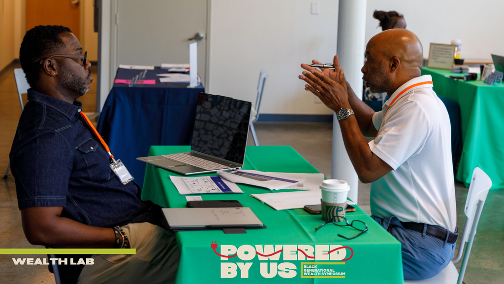 Two men sit across a table at a business event, one gesturing while speaking. Laptops, paperwork, and a coffee cup are on the table. WEALTH LAB and POWERED BY US text is visible on the image, highlighting themes of legacy and Black philanthropy as part of Black Philanthropy Month.