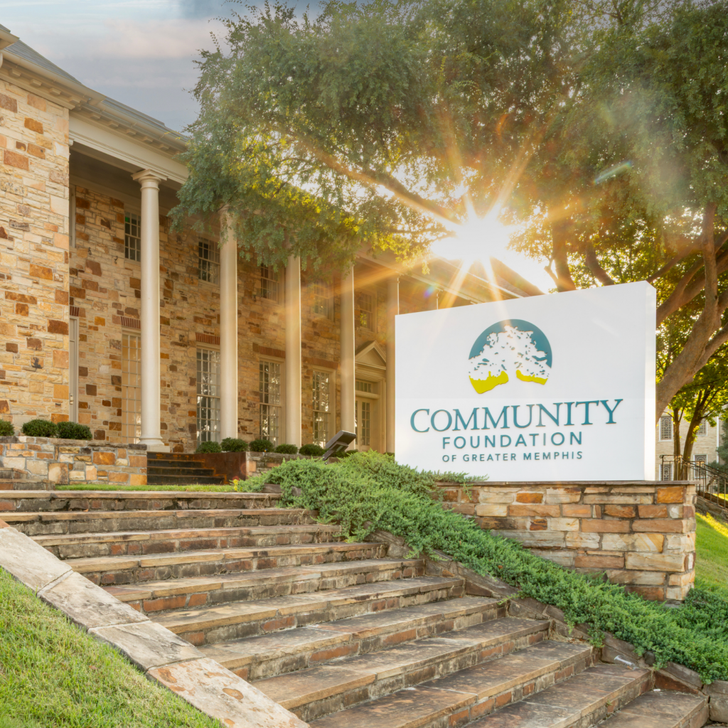 A stone building with columns is seen at sunset. A sign in front reads Community Foundation of Greater Memphis. Steps and green bushes lead up to the entrance.