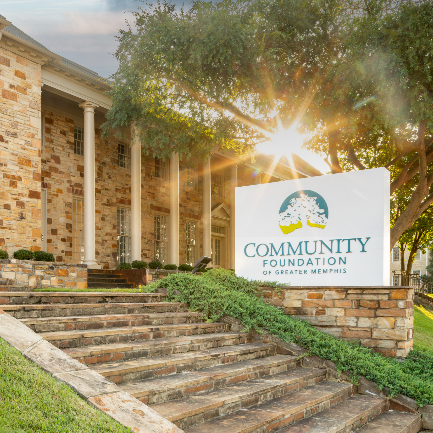 A stone building with columns is seen at sunset. A sign in front reads Community Foundation of Greater Memphis. Steps and green bushes lead up to the entrance.