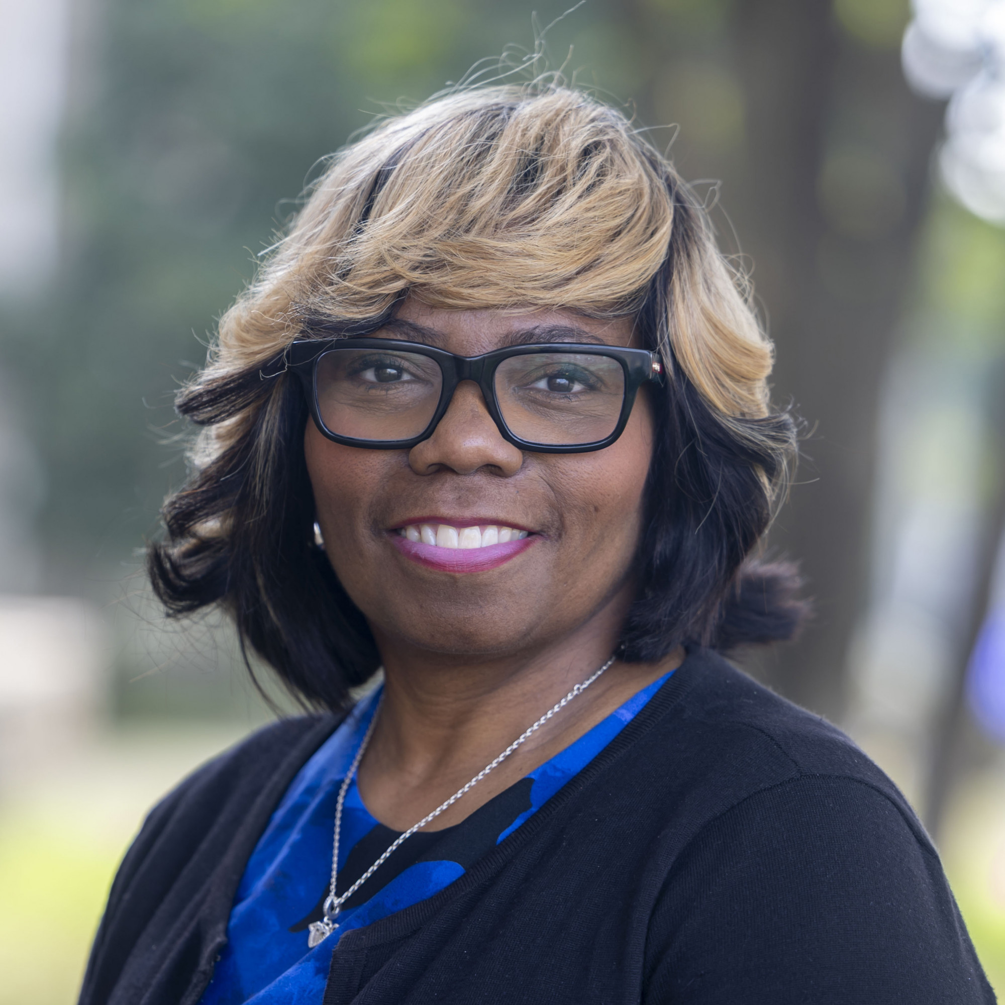 Yvette Washington, a woman with shoulder-length black and blonde hair, wearing glasses, a blue top, and a black sweater, smiles outdoors with trees and greenery blurred in the background.