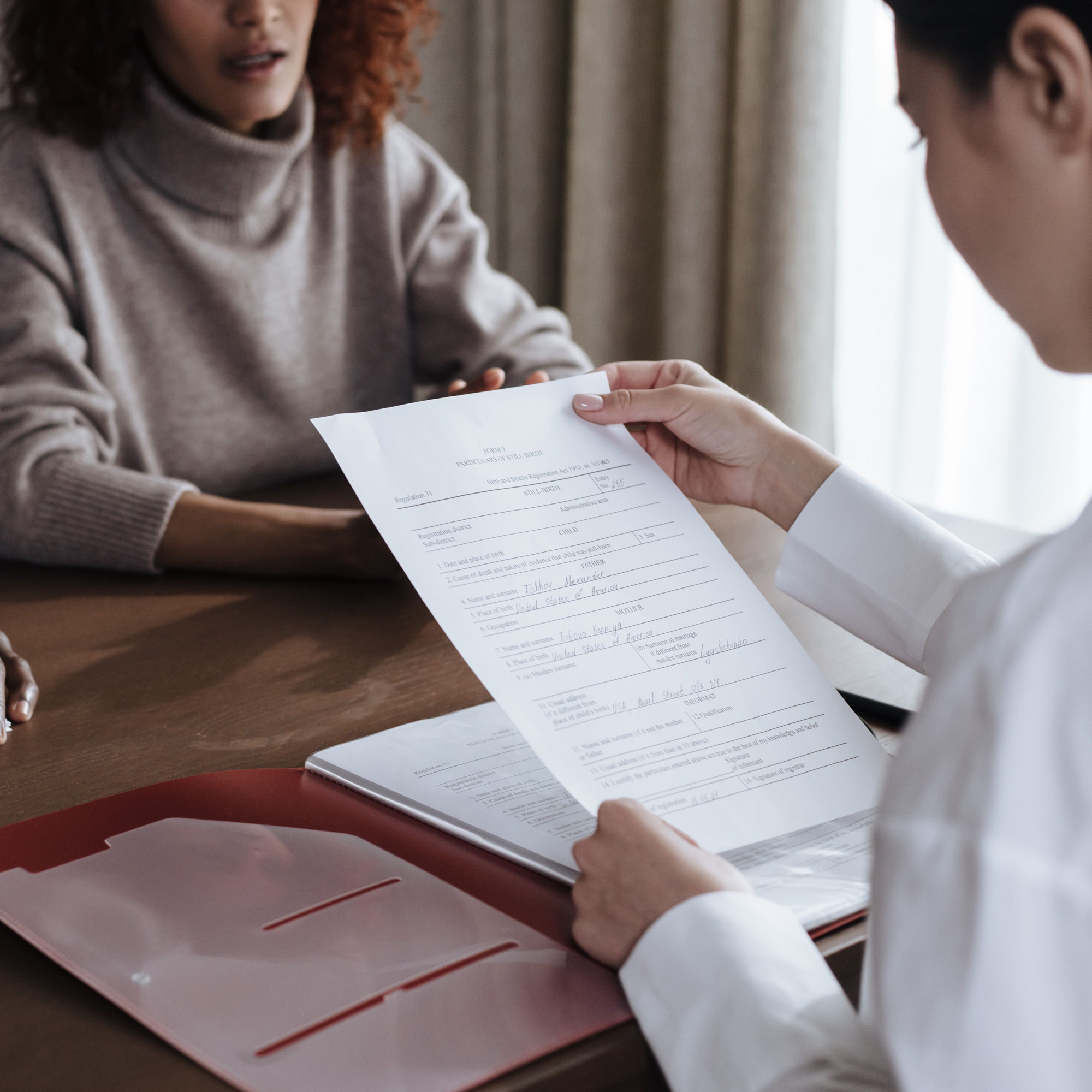 A person in a white shirt holds and reviews a printed resume while sitting across a table from another person in a beige sweater, perhaps discussing topics like estate planning or the importance of including the 13 magic words. A red folder lies open on the table.