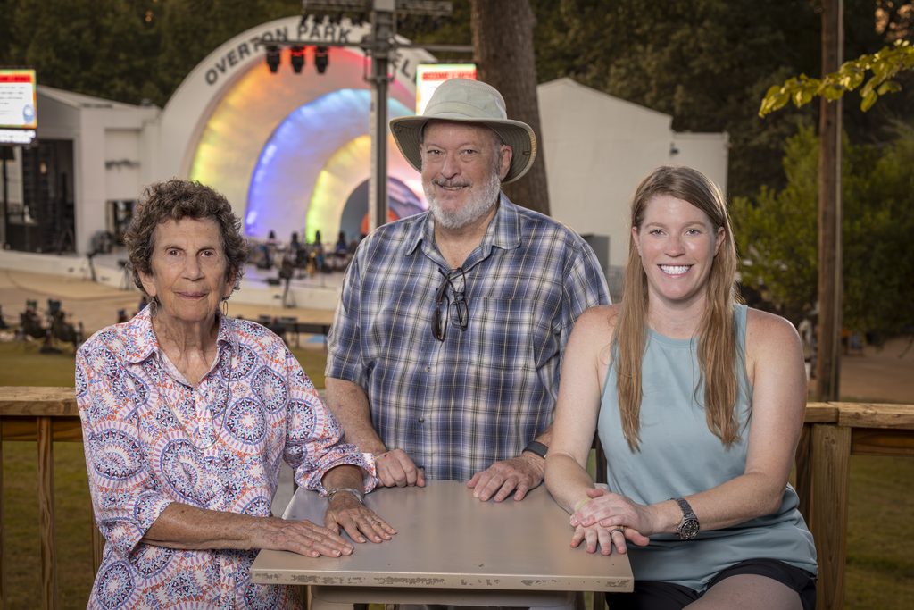 Three people, two women and one man, smile while sitting at a table outdoors. An amphitheater with a colorful lit arch, trees, and people in the background suggest a park or public event setting rooted in Memphis.