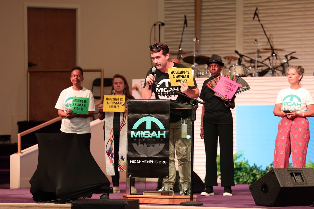 A man speaks at a podium labeled MICAH, emphasizing the importance of the long game in advocating for change, while others stand behind him holding signs that read Housing is a Human Right! in a church setting.