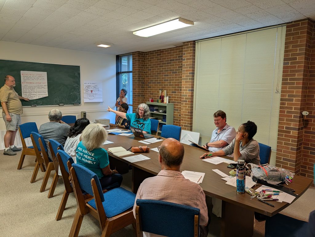 A group of people sit around a table in a meeting room, clearly focused on discussing their long game strategy. One person stands by a chalkboard with notes, while another points towards the front. Papers and notebooks are spread on the table.
