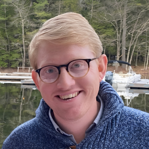 A smiling man with blonde hair and glasses wearing a blue knit sweater, with a lake and boats in the background.
